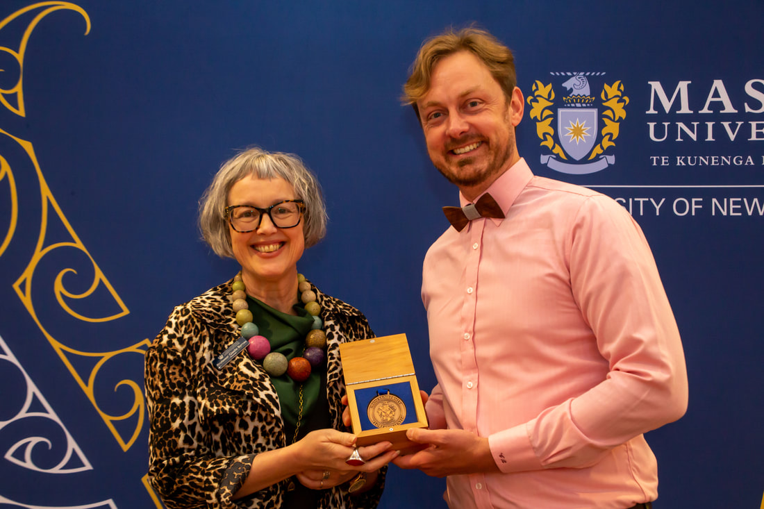 Collin Bjork, a white man in a pink dress shirt holds a wooden box with a brass teaching medal alongside a white woman, Provost Giselle Byrnes, in a multicoloured necklace. Both are smiling in front of a blue Massey University sign at a teaching and research excellence ceremony.