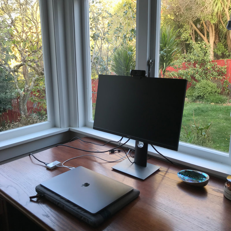Laptop and monitor on an oak desk in front of a double window looking out at a yard.