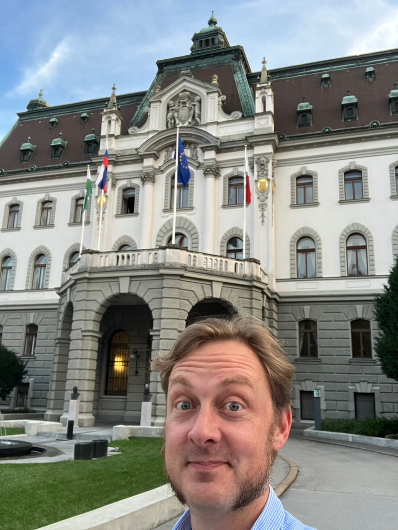 Smiling white man with long sideburns in a collared shirt outside of a tall regal looking austro-hungarian building that is the main building of the University of Ljubljana.