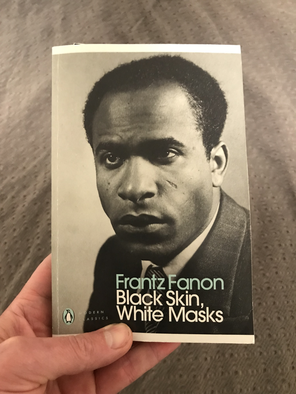 White fingers of Collin Bjork holding Frantz Fanon's Black Skin, White Masks book, which has a black and white portrait of Fanon on the front