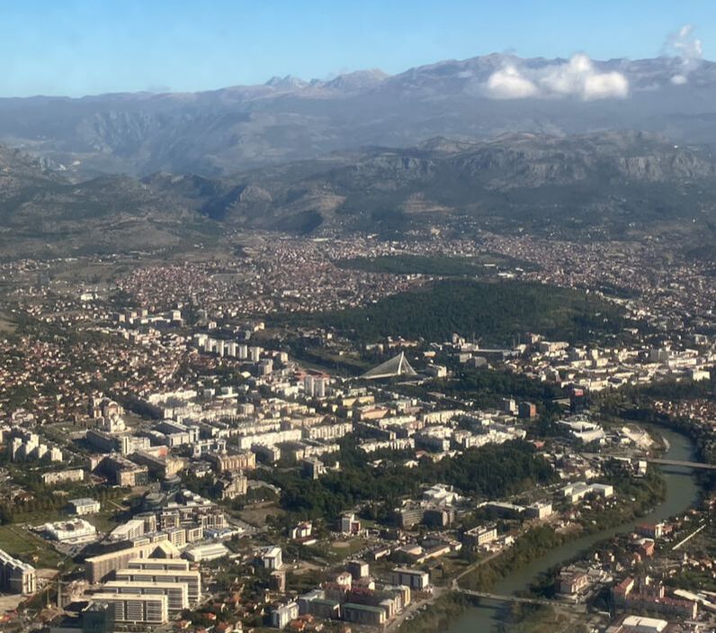 Photo of Podgorica from an airplane window, a modern city with a river, white buildings, and mountains. Taken by Collin Bjork